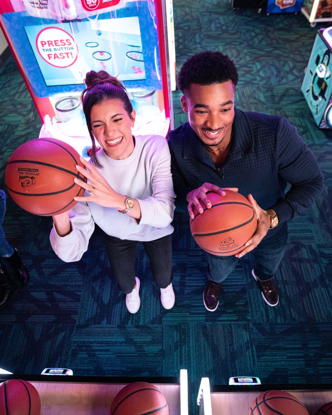 photo from a high angle looking down at a woman and a man each holding a basketball. Looks like they are about to throw the basketballs. behind them is an arcade game with white and red lights.
