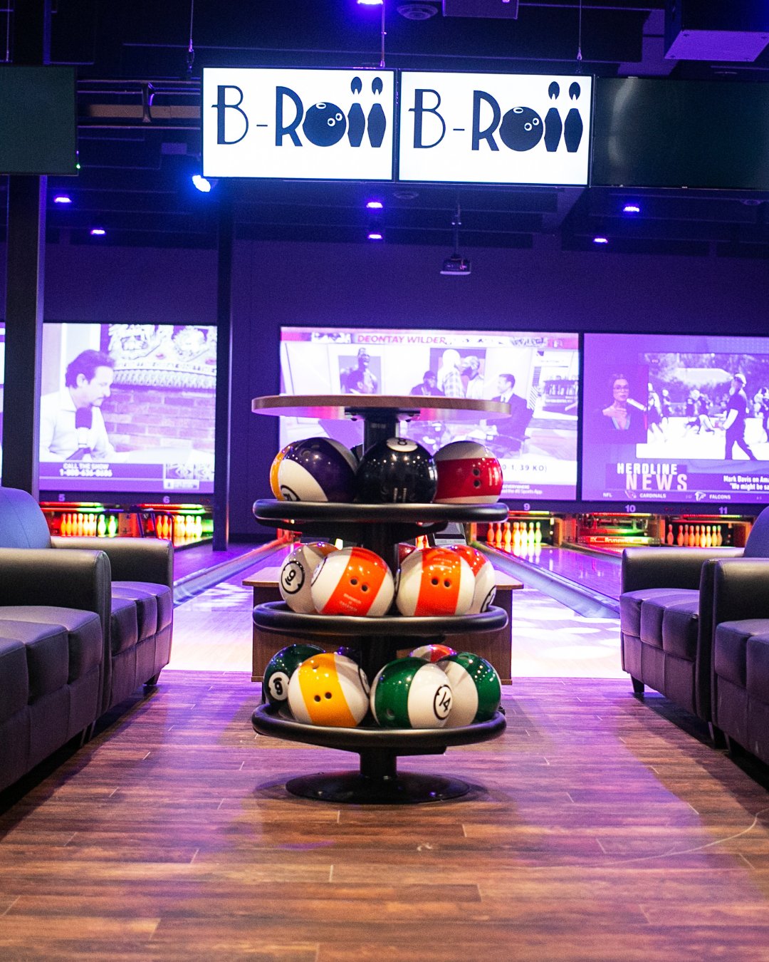 bowling ball stand in the center. in the background bowling lanes & tvs. surrounded by seating on the outer edges