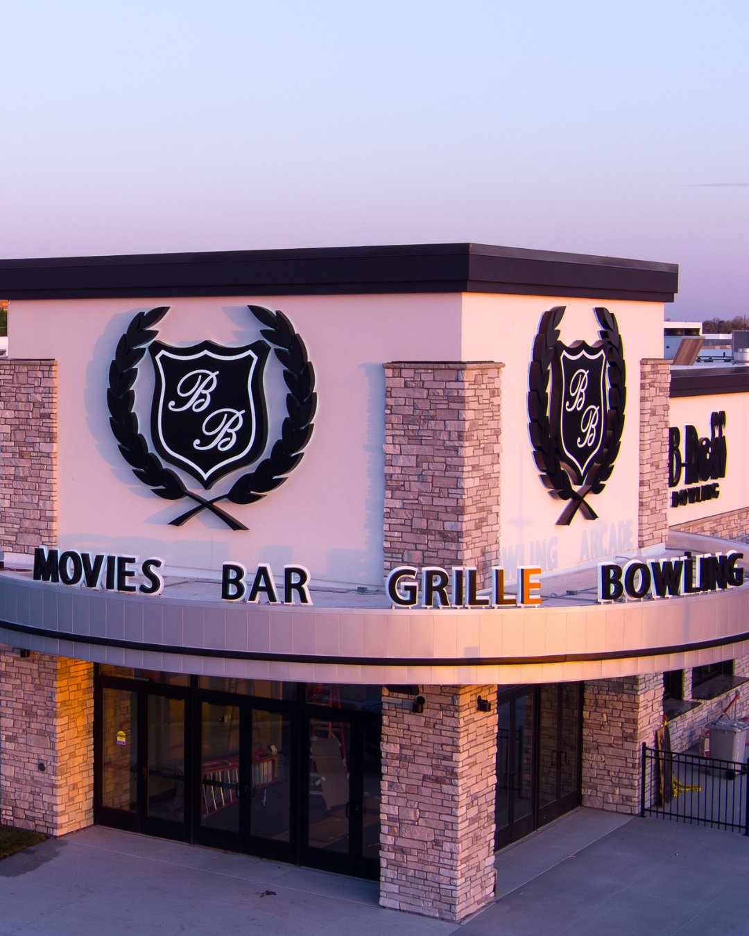 exterior of building entrance. A semi-circle around the corner of the building has signage that reads "MOVIES BAR GRILLE BOWLING" On 2 sides of the building are 2 giant B&B Crests