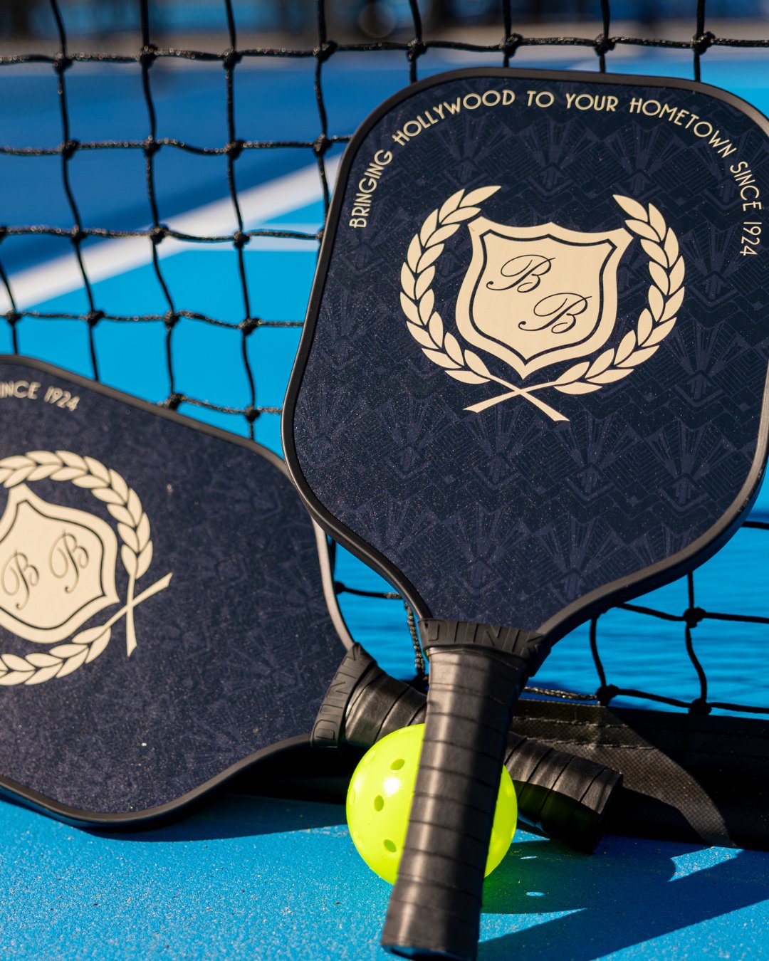 blue pickleball surface. 2 black pickleball paddles with B&B logo leaned up against the black net. 1 bright neon yellow pickleball