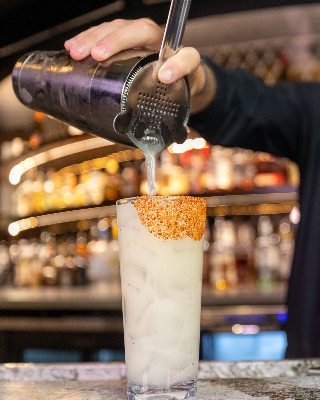 tall glass filled with margarita & ice, rim lined with tajin. Bartender's arm holding a shaker & pouring the drink into the glass