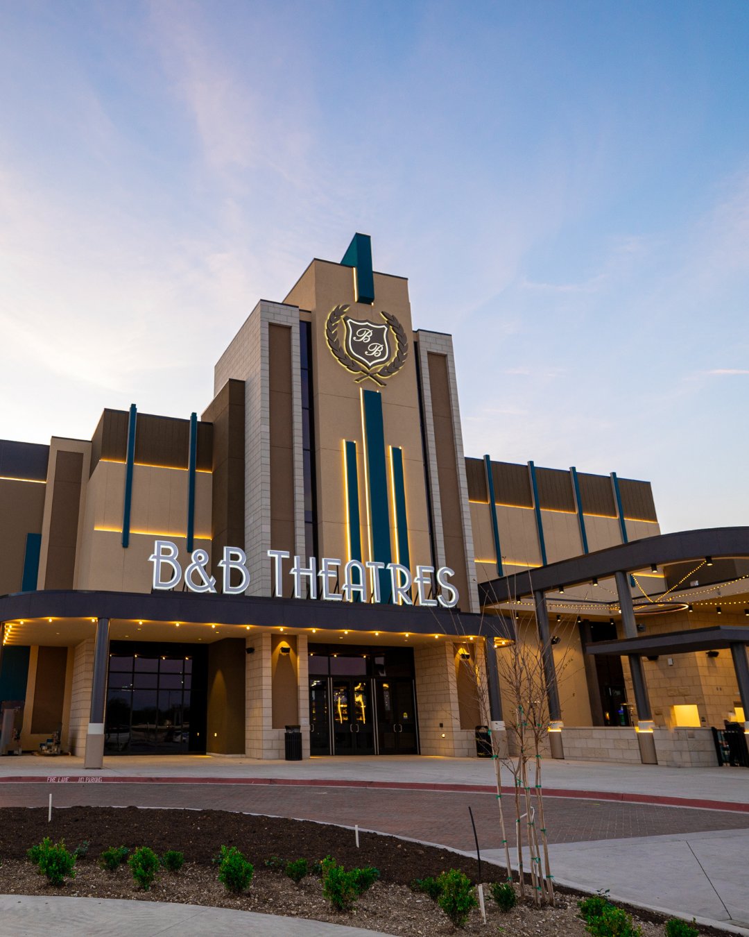 exterior of the building at sunset. Lit white signs read "B&B Theatres" there is also a lit up crest near the peak of the building