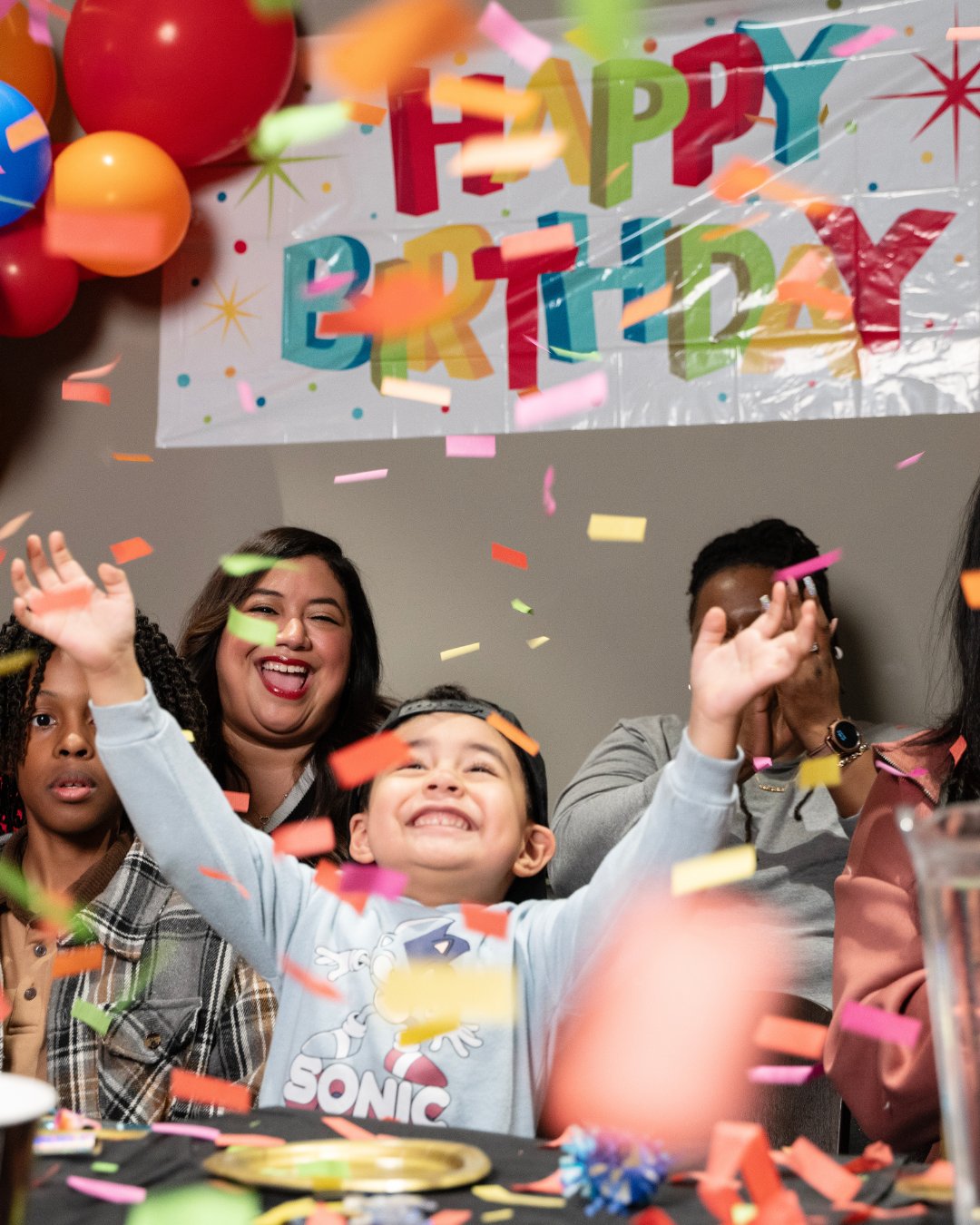 close up of a child with their arms raised looking up at colorful confetti falling around him. behind him is 1 child and 2 adults smiling & clapping. There is a white banner taped to the wall with happy birthday written in colorful letters.