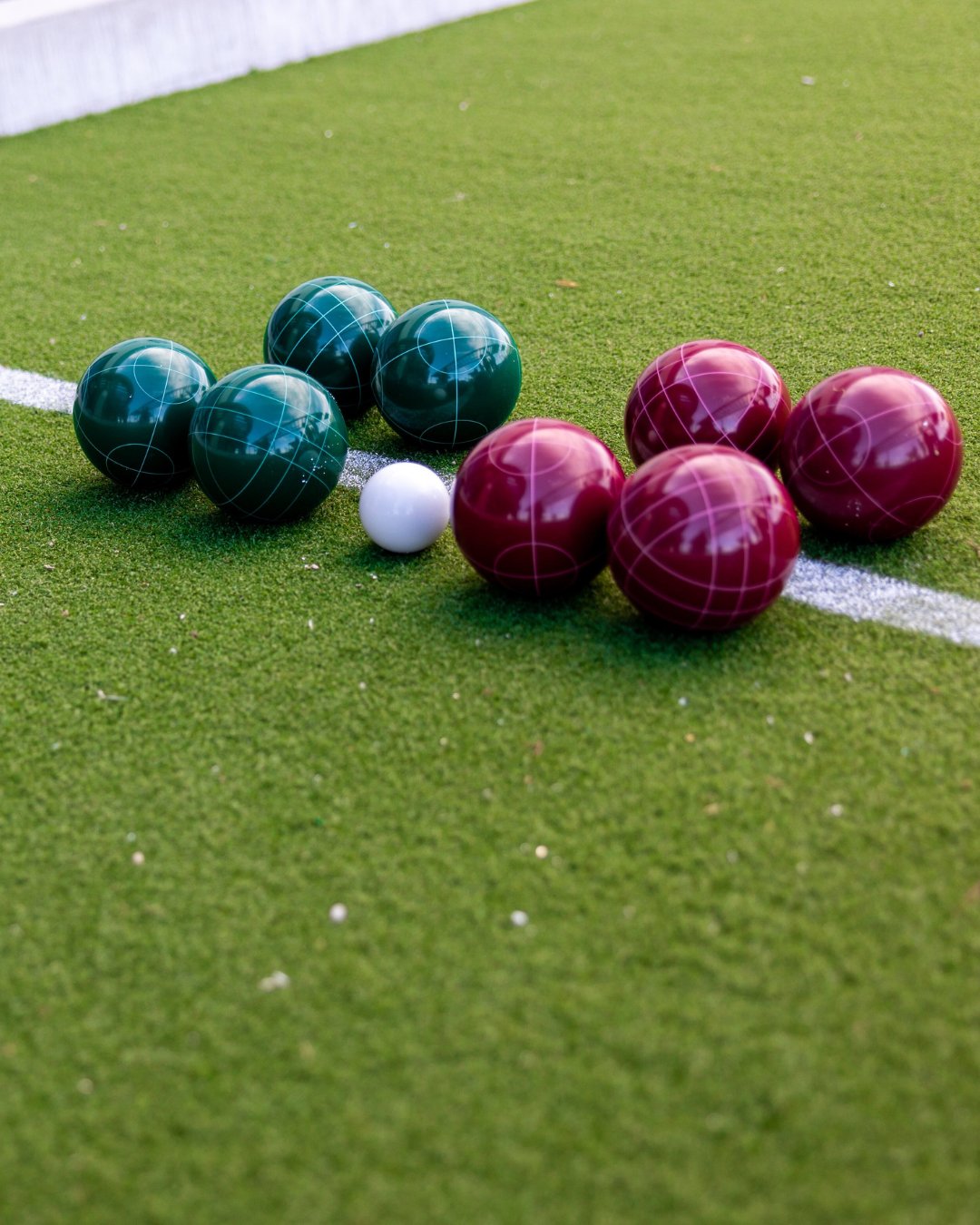 green turf with a white line horizontally down the center. sitting near the white line are 4 green balls, 4 maroon colored balls, and 1 smaller white ball.