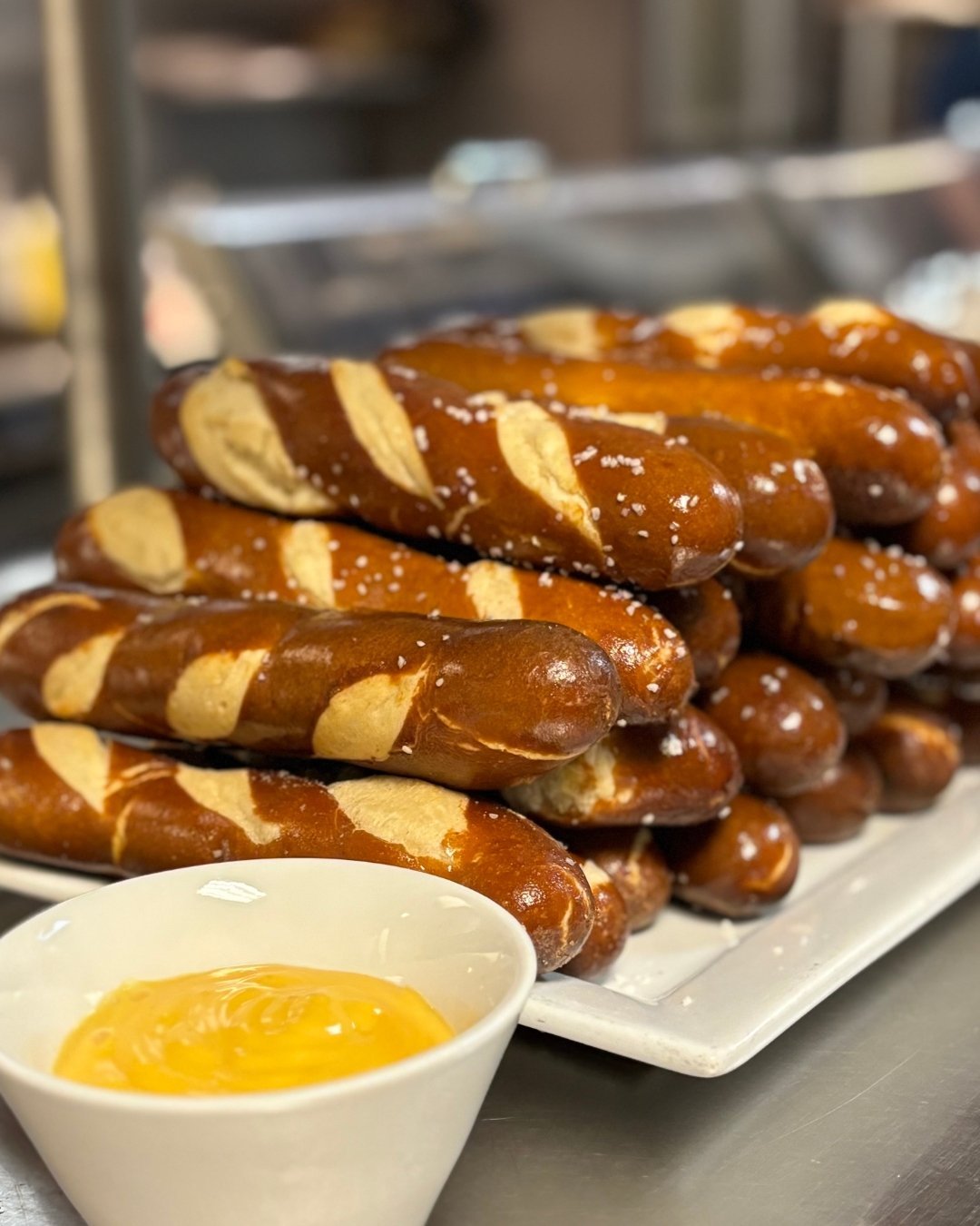 rectangular plate filled with stacked pretzel sticks. The pretzels are topped with salt. In the foreground is a small bowl of cheese dip