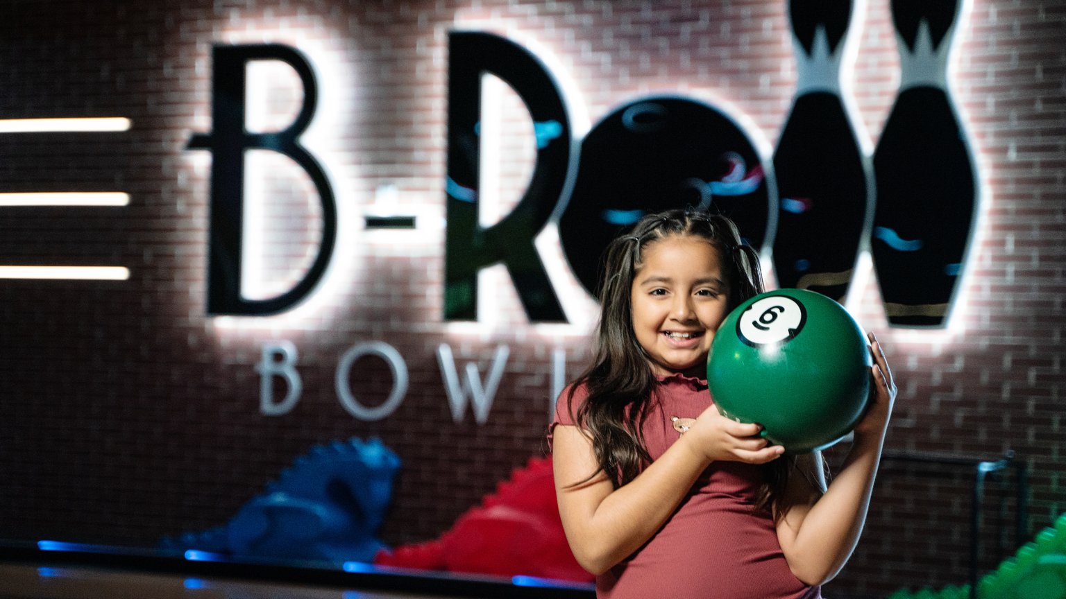 young girl standing and holding a green bowling ball, she's smiling. Behind her is a lit up sign that reads B-Roll Bowling