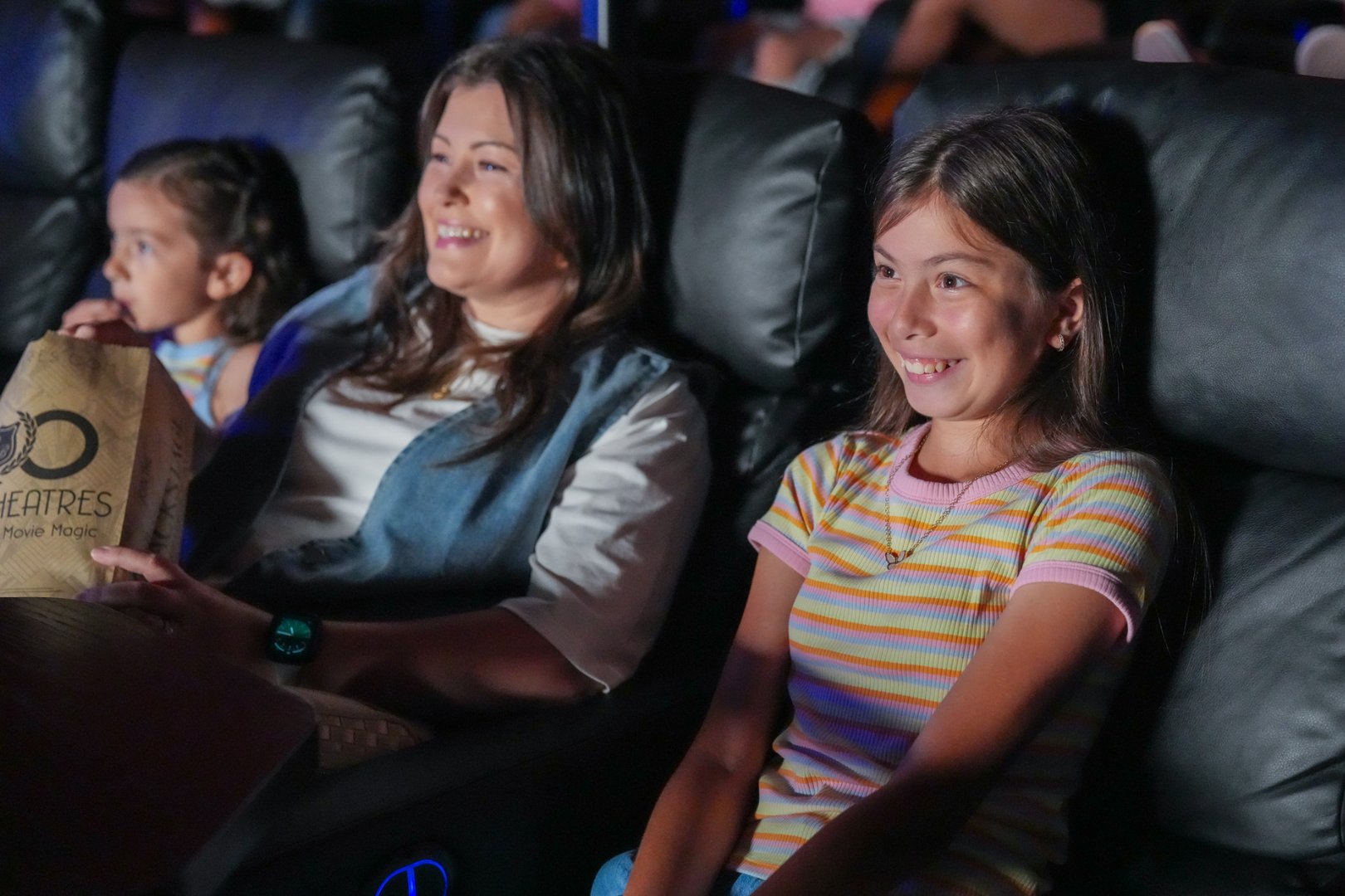 a woman sitting between two young kids smiling at a movie screen (out of frame) the woman is holding a bag of popcorn