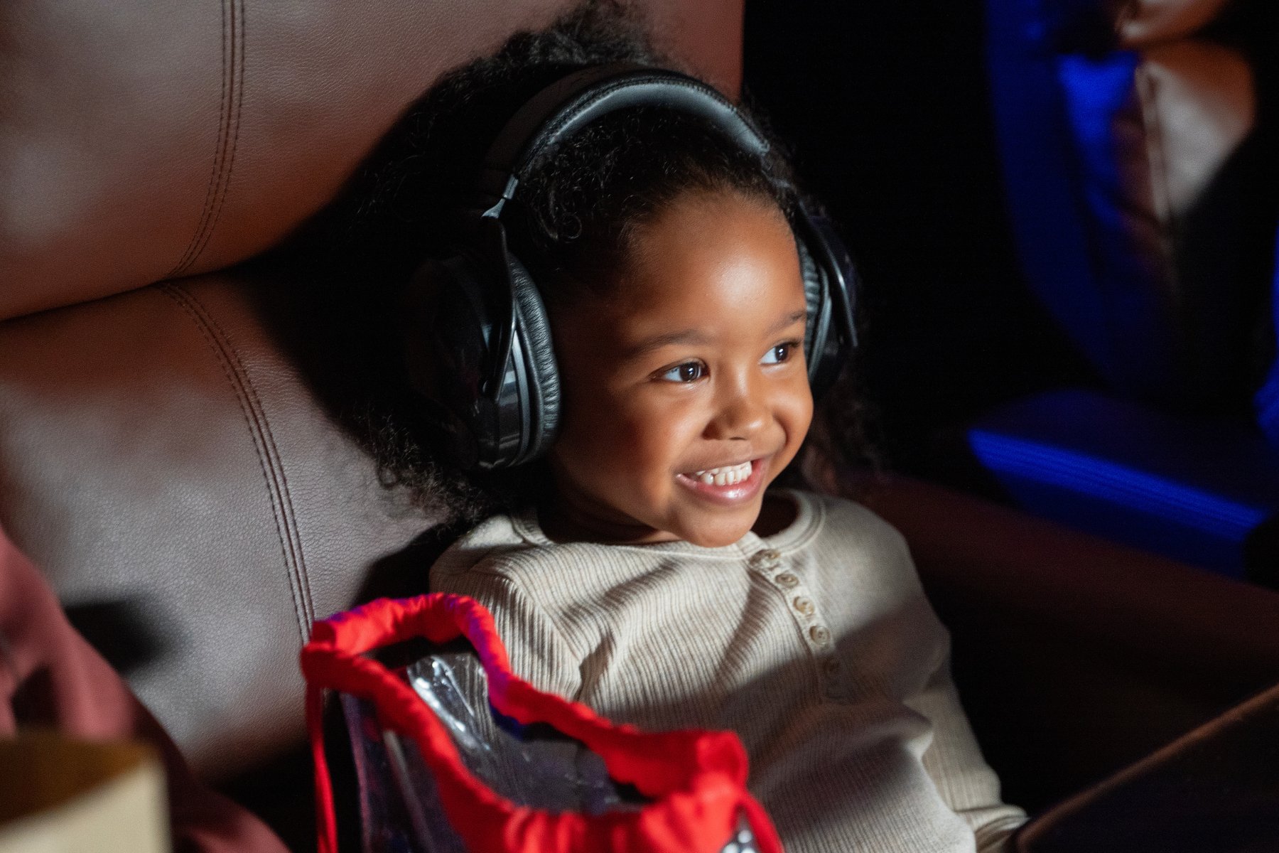 small child sitting in a recliner. Close up on her face, she's smiling & wearing noise cancelling headphones. Next to her in the seat is a bag with a red cinch drawstring closure.
