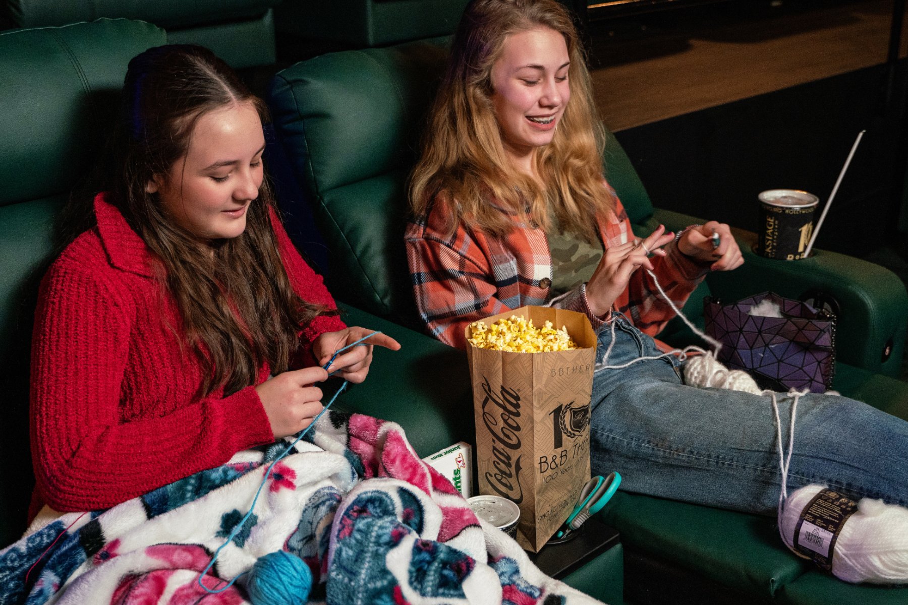 2 young women sitting in green recliners. They have crochet hooks and yarn in their laps. There is a large bag of popcorn in between them.