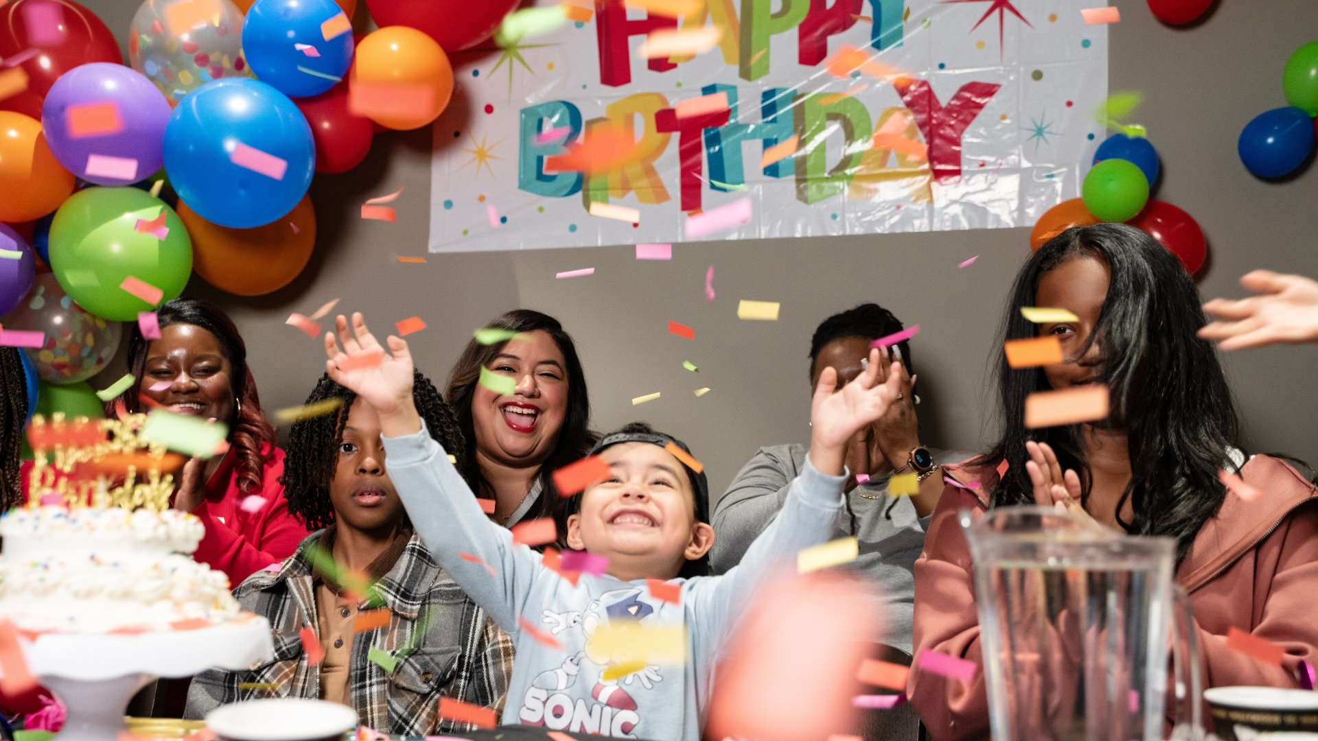 close up of a child with their arms raised looking up at colorful confetti falling around him. surrounding him is 2 children and 3 adults smiling & clapping. There is a white banner taped to the wall with happy birthday written in colorful letters.