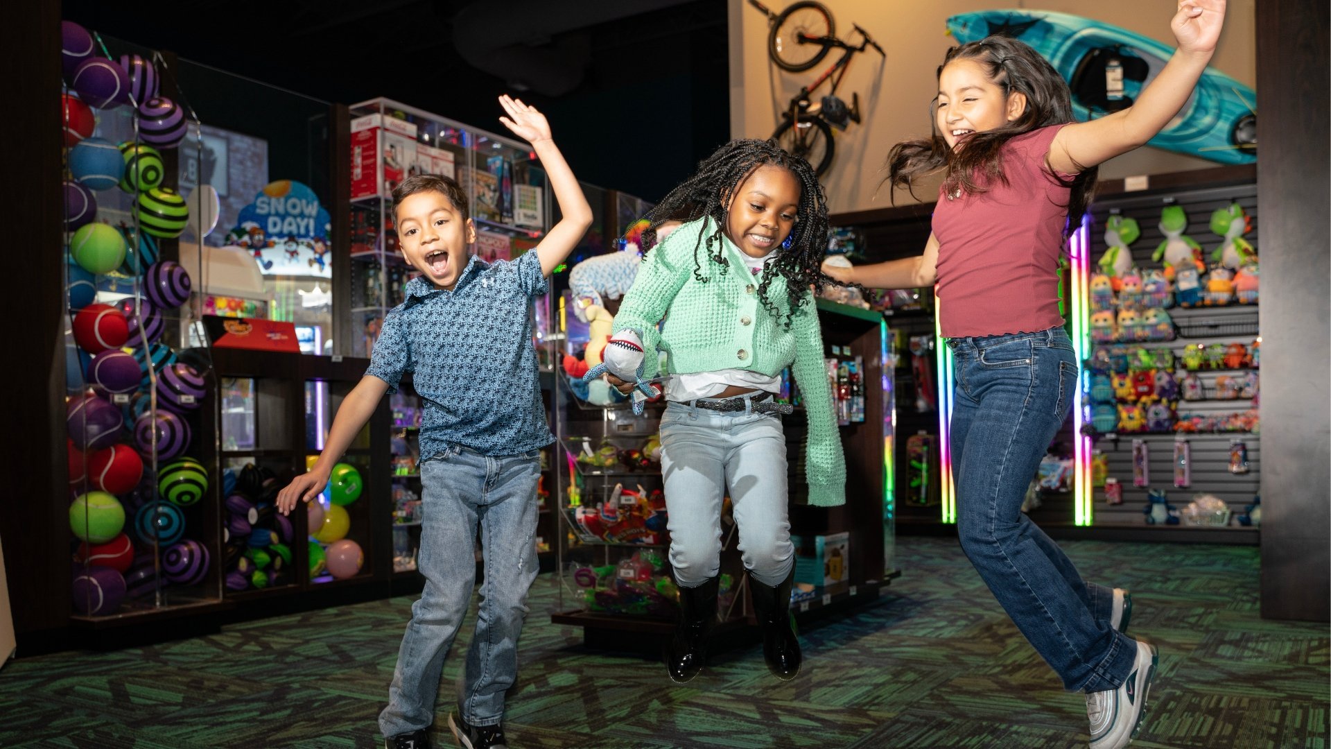 3 children are jumping in the air with big smiles on their faces and hands raised in the air. They are photographed in front of the arcade redemption store
