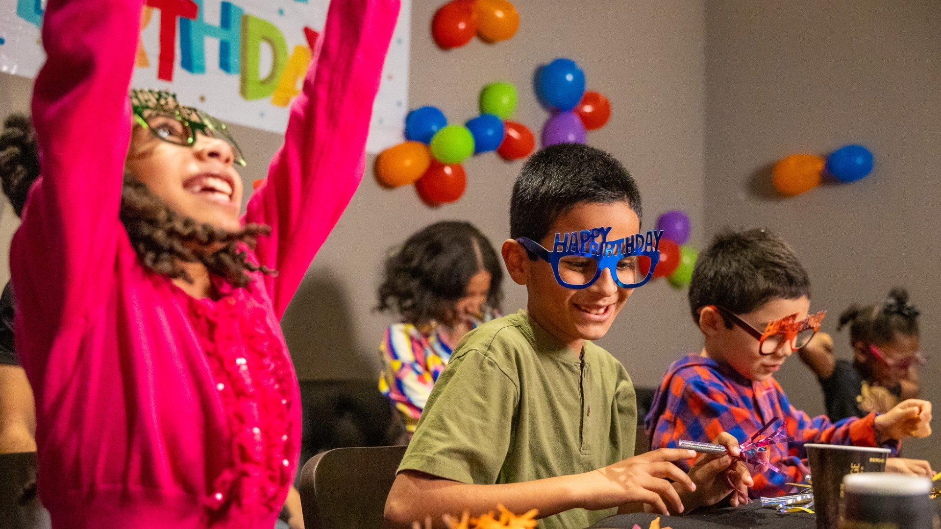 3 kids with birthday glasses on smiling & celebrating. room is filled with colorful balloons taped to the walls.
