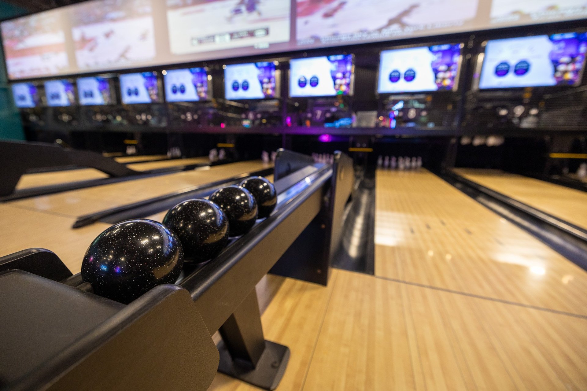 image standing behind the ball return facing the pins. In the foreground 4 balls resting in the ball return, in the background many TVs above the duckpin pins.