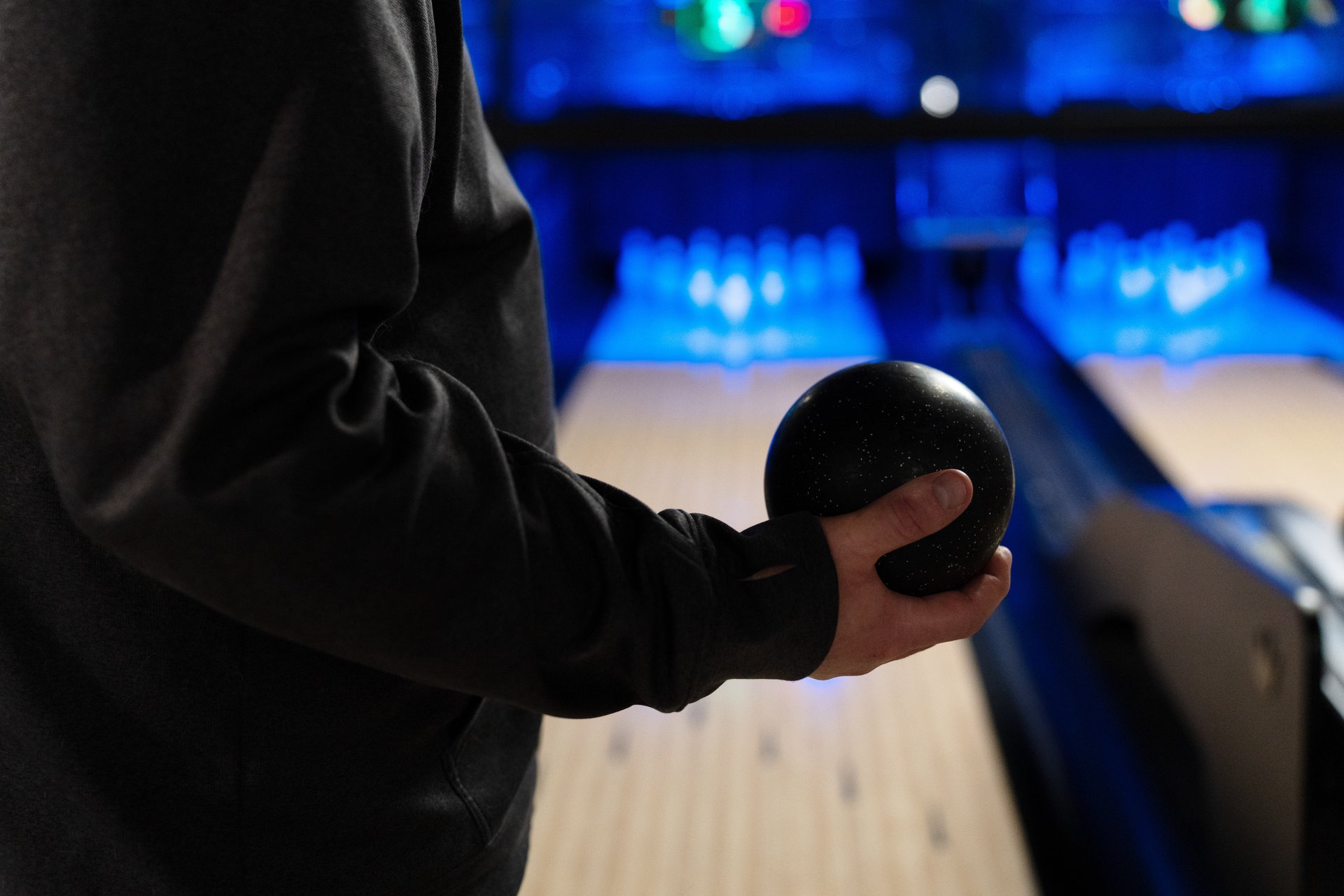 person wearing black long sleeve jacket standing perpendicular to camera and taking up left side of image. They are holding a small black duckpin ball in their right hand. Lane and pins in the background