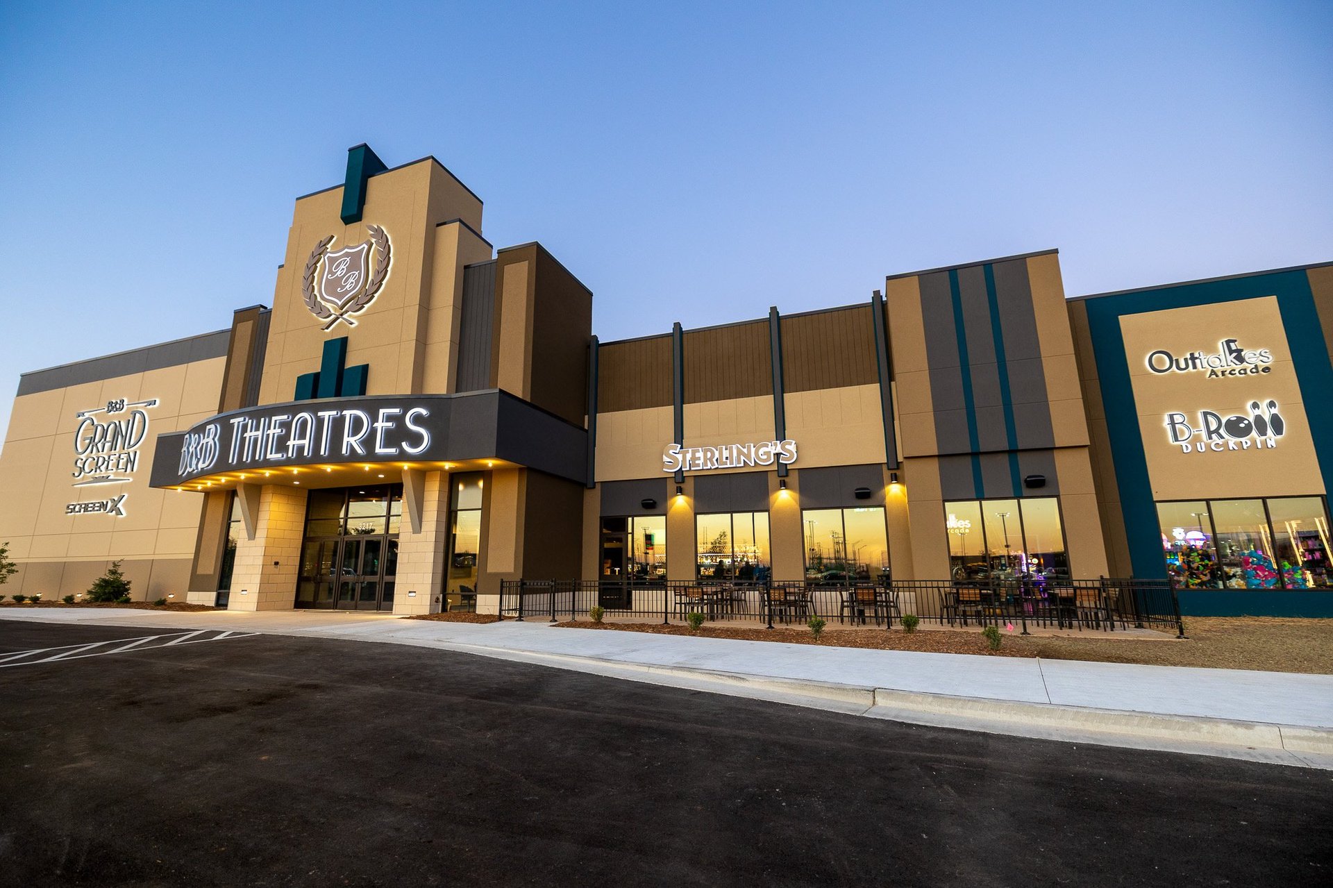 external view of the building at sunset. Building has signage that is lit up. Signs say B&B Theatres, Grand Screen SCREENX, Outtakes Arcade, B-Roll Duckpin, Sterling’s