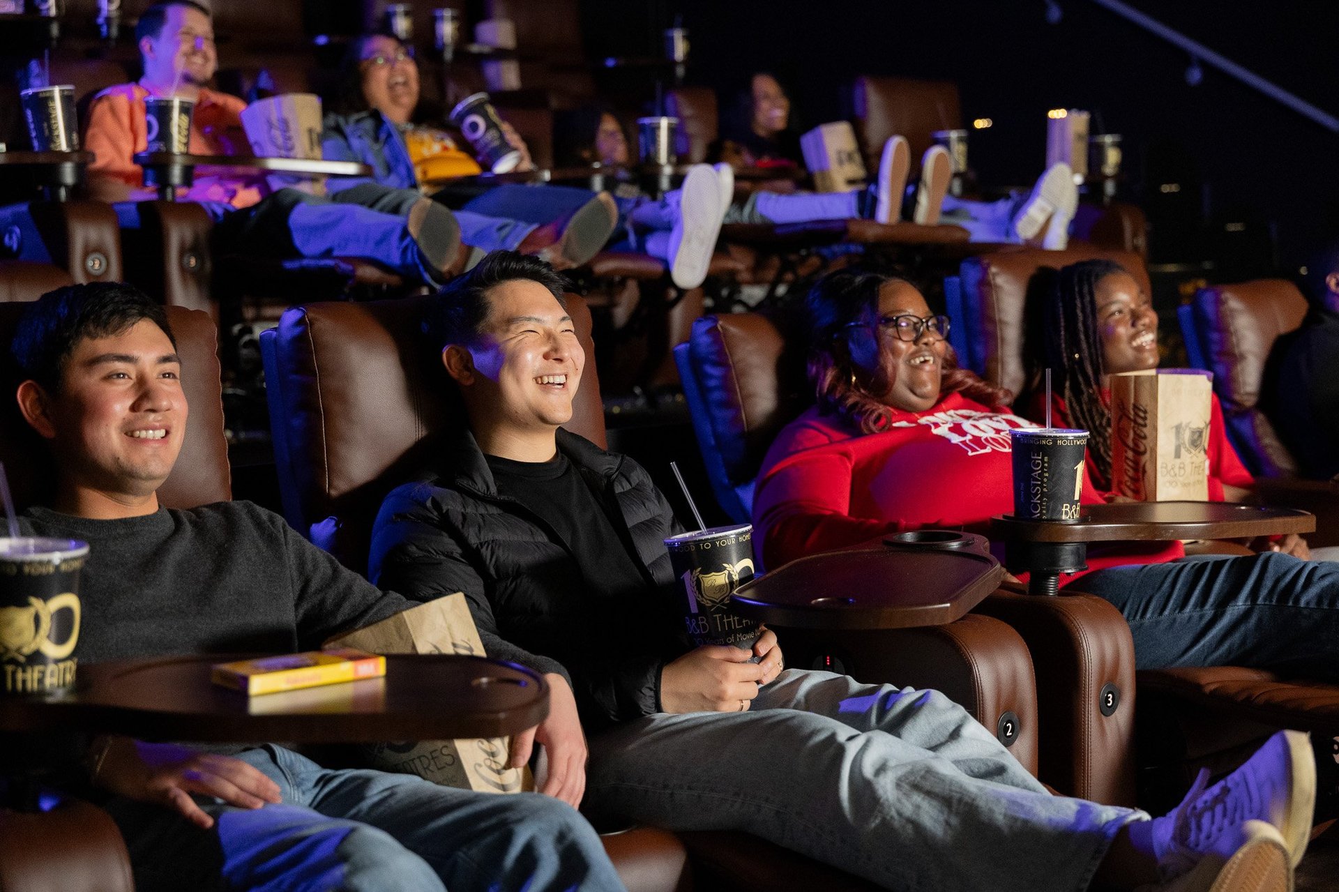 2 rows full of guests. All are smiling. Some are holding popcorn bags, others cups. They are looking toward a movie screen (out of frame)