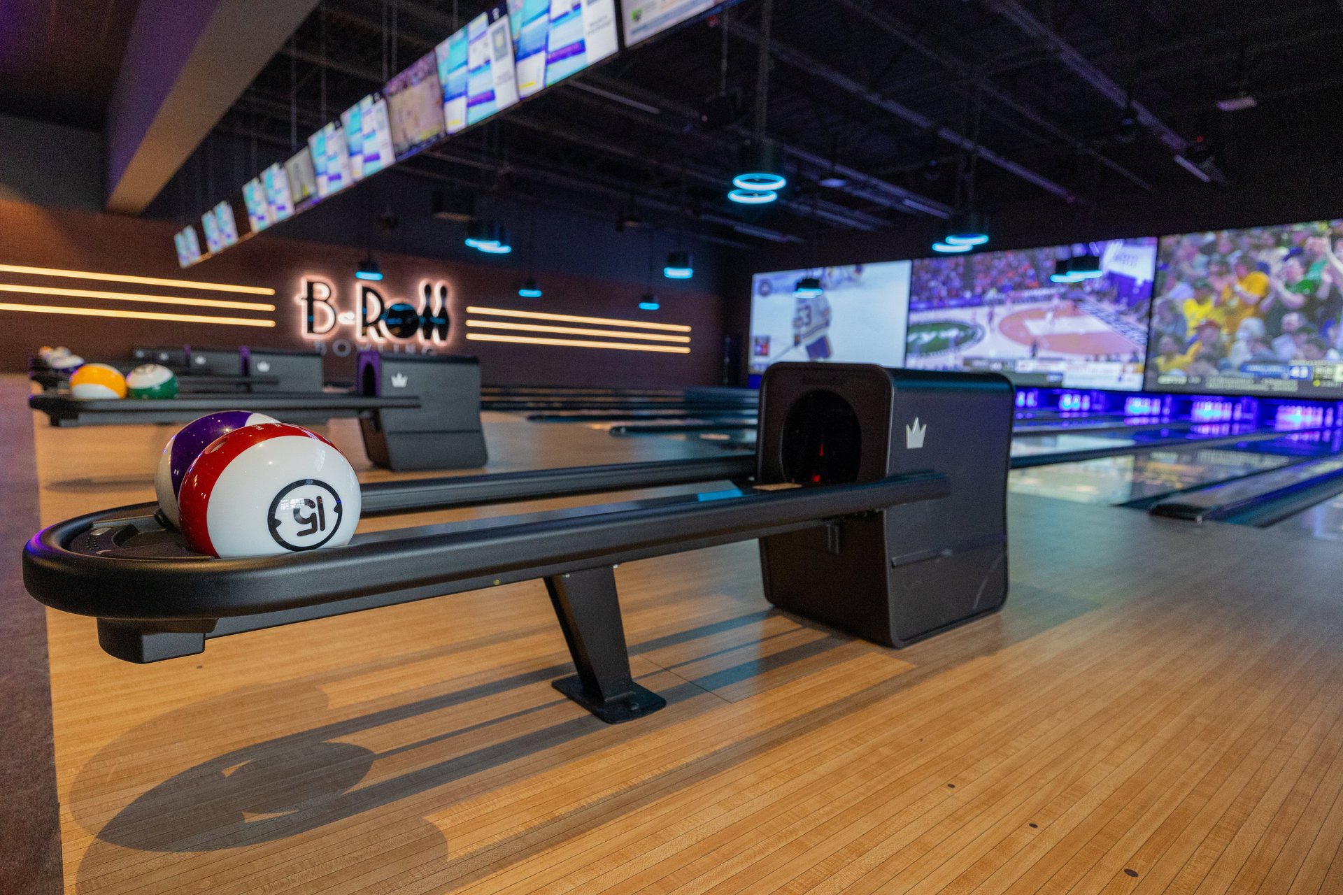 Bowling ball return with 2 balls sitting in the return. 1 is white with a red stripe and has the number 15 written in the center. In the background are bowling lanes, on the back wall is a lit sign that reads B-Roll