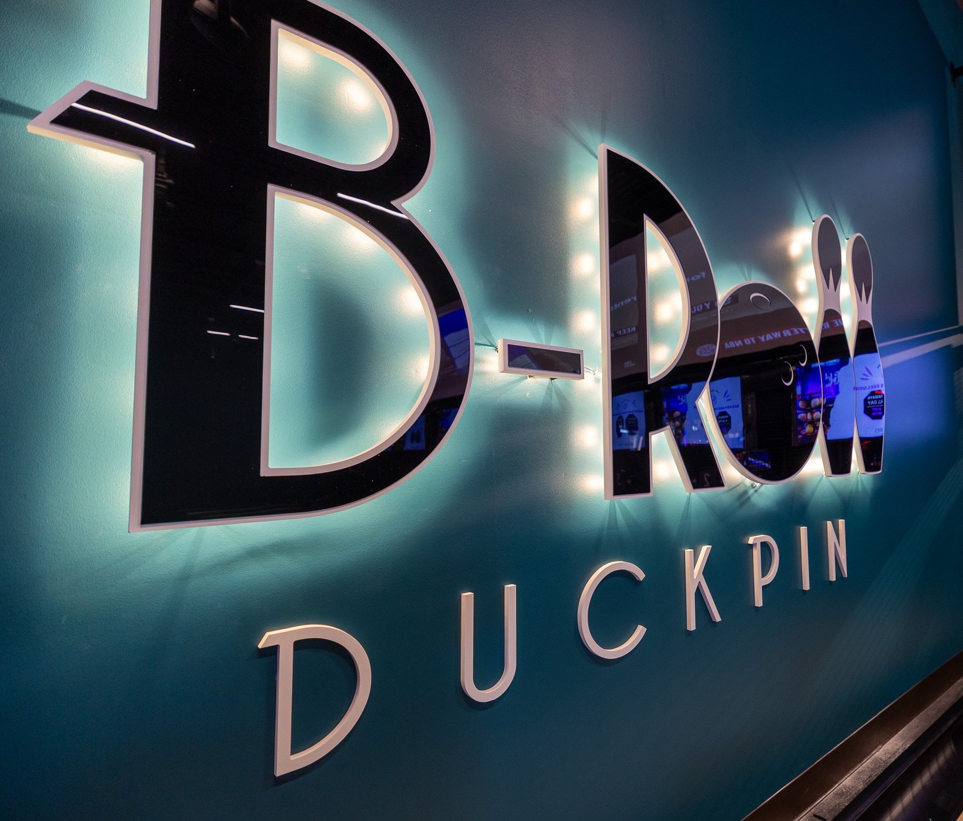 blue-ish wall with 3d lit-up signage that reads B-Roll Duckpin. B-Rill is large and black with a bowling ball for the letter 'o.' Duckpin is underneath, is smaller, and white font