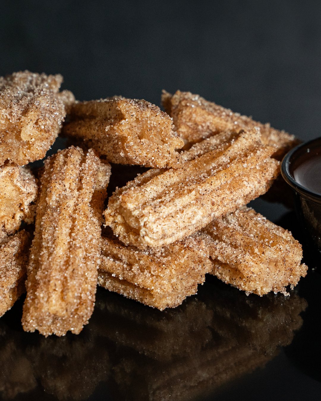 close up image of pile of churros with a small circular container of chocolate dipping sauce to the right. 