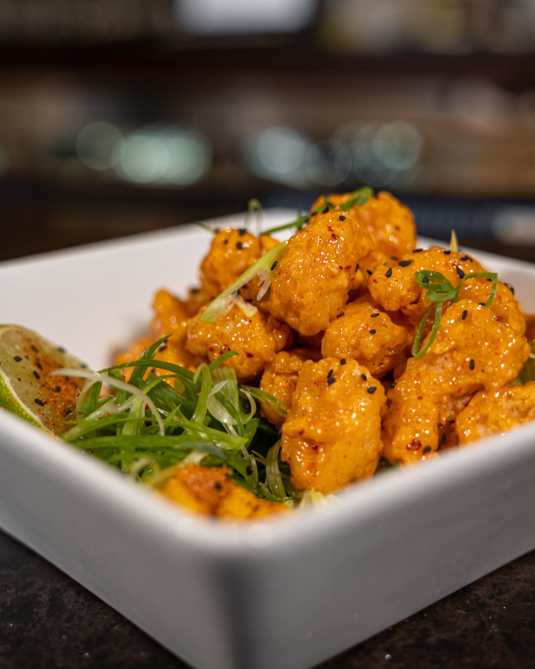 close up of a white, square bowl filled with chicken bites, topped with sesame seeds, greens, and a lime wedge in the corner