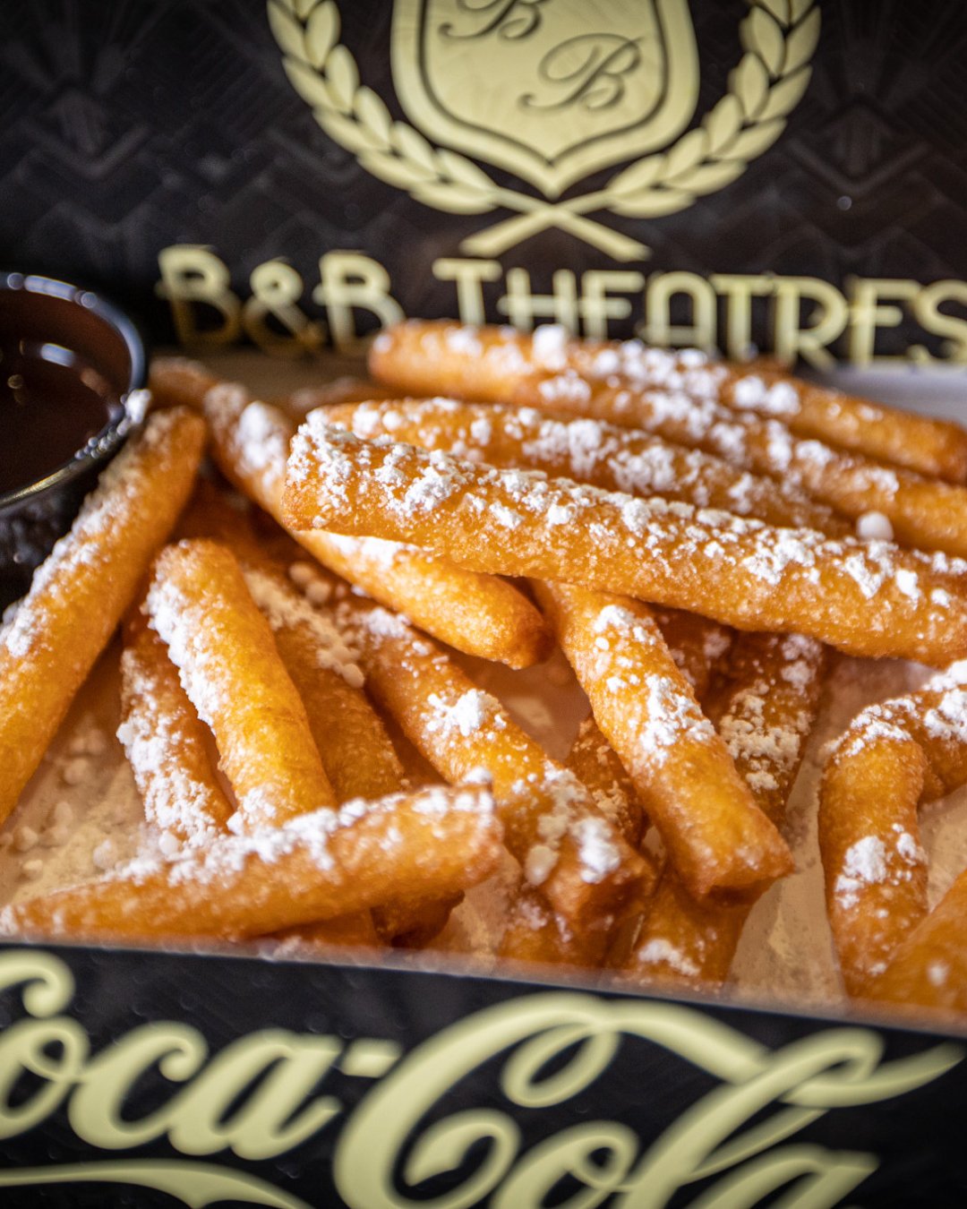 close up image of pile of fried funnel cake fries with a small circular container of chocolate dipping sauce to the left. 