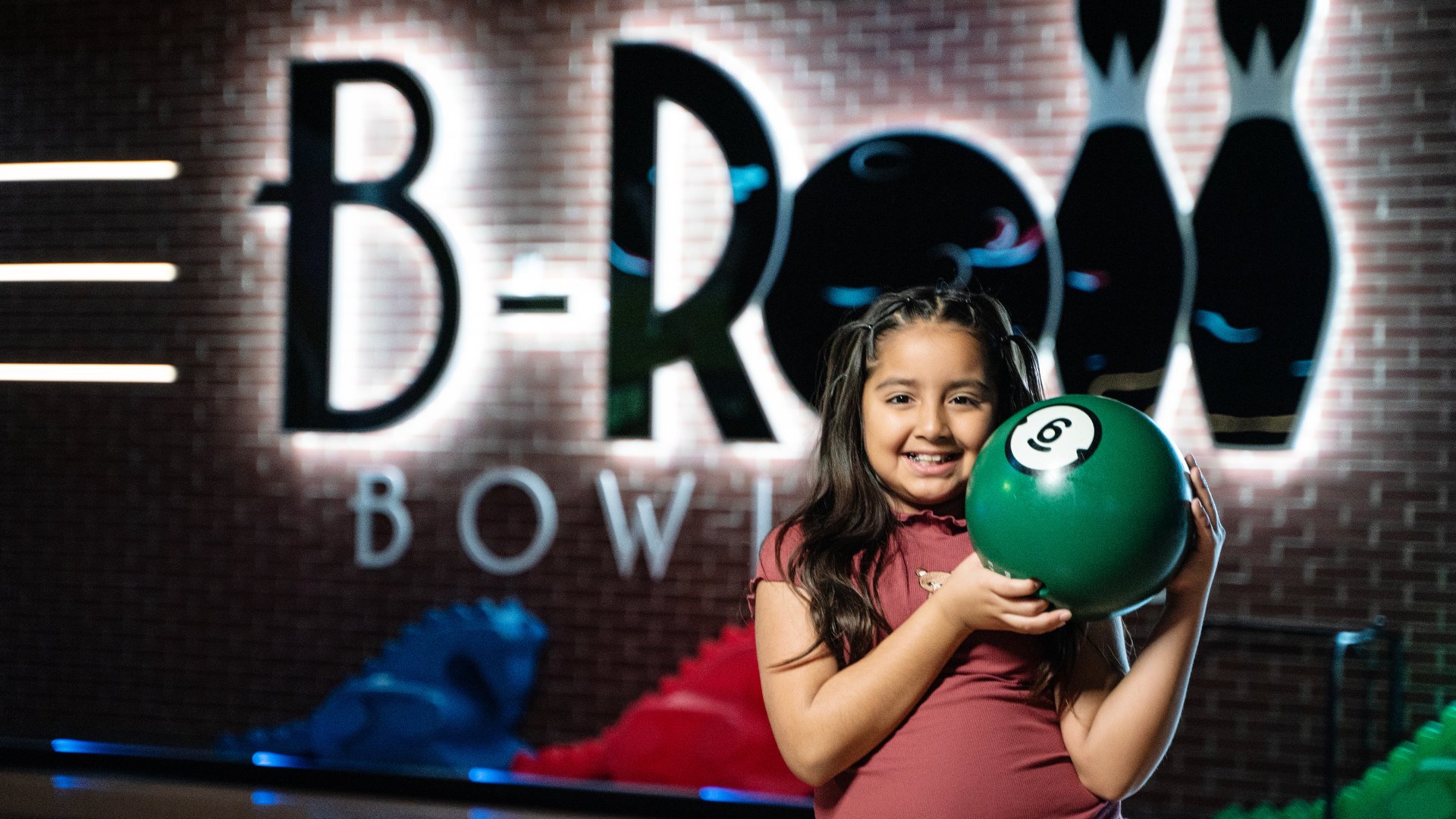 young girl holding a green bowling ball next to her face & smiling. In the background is a giant lit up sign that reads B-Roll BOWLING