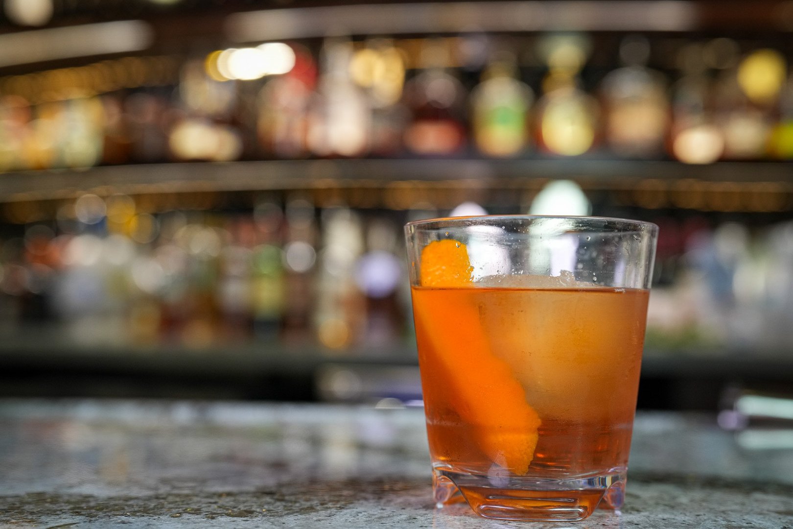 old fashioned sitting on a bar top filling the right side of the frame. There is a big orange peel sitting in the cocktail