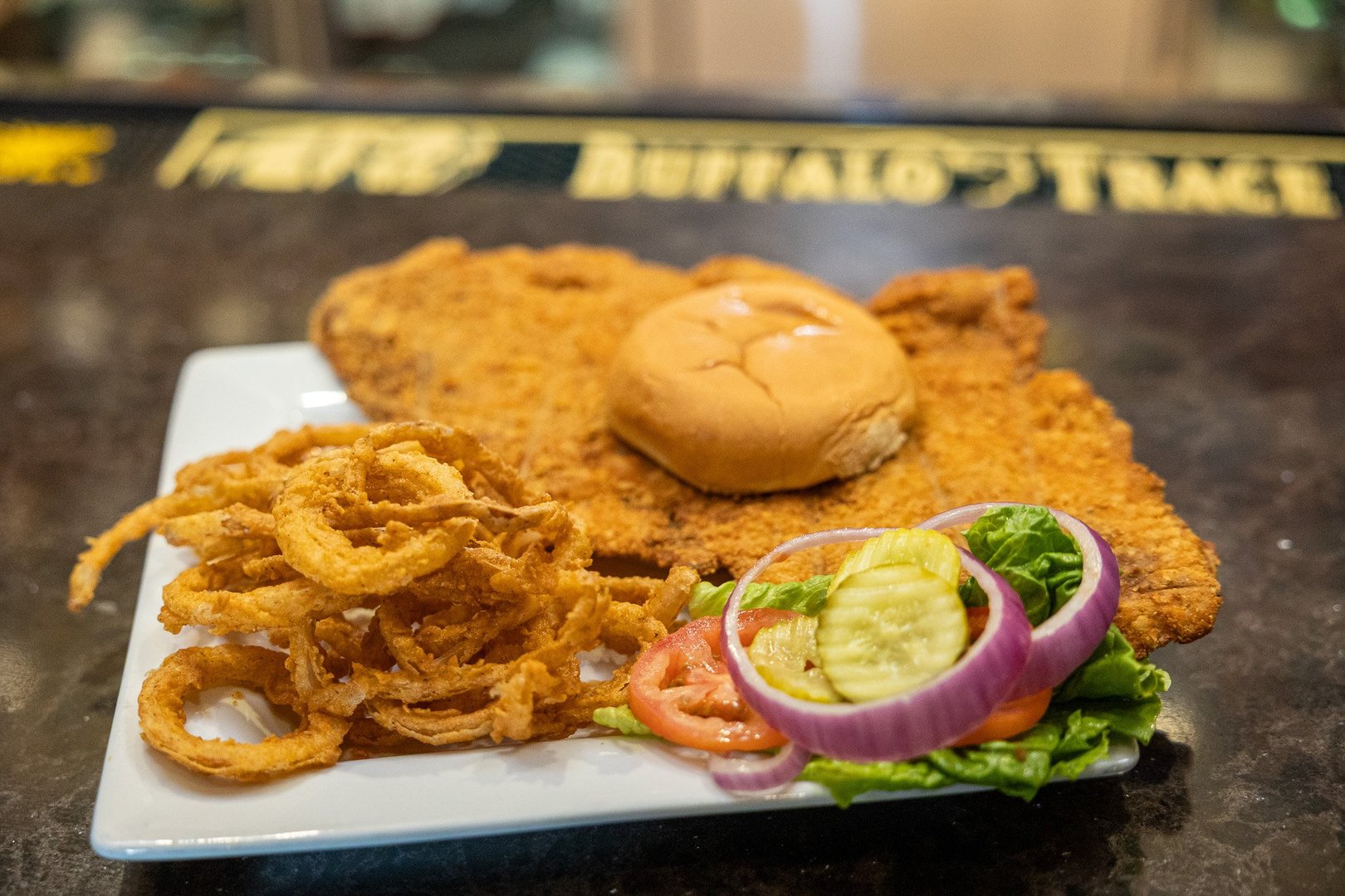 square white plate topped with a giant pork tenderloin (so big it's hanging passed the edge of the plate) with a tiny bun placed on top. Surrounding the sandwich are fried onion strings & lettuce, tomato, onion, pickle accoutrements