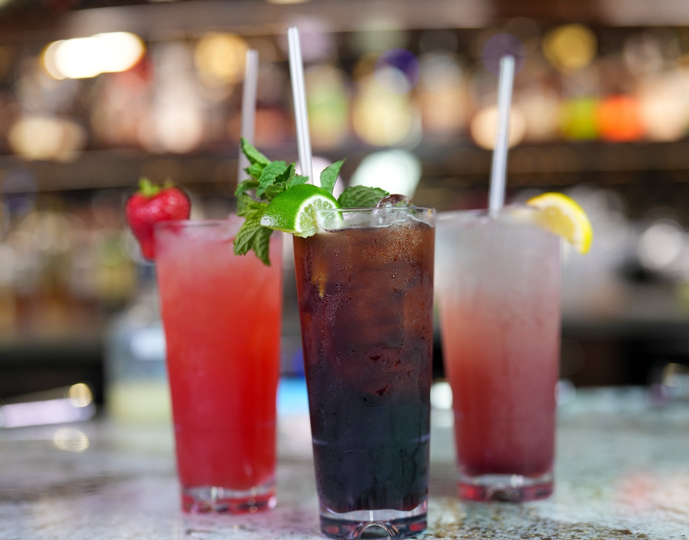 3 tall glasses sitting next to each other on a bar. From left to right: Sparkling Lemon Strawberry, Raven's Cauldron, Lemon Blueberry