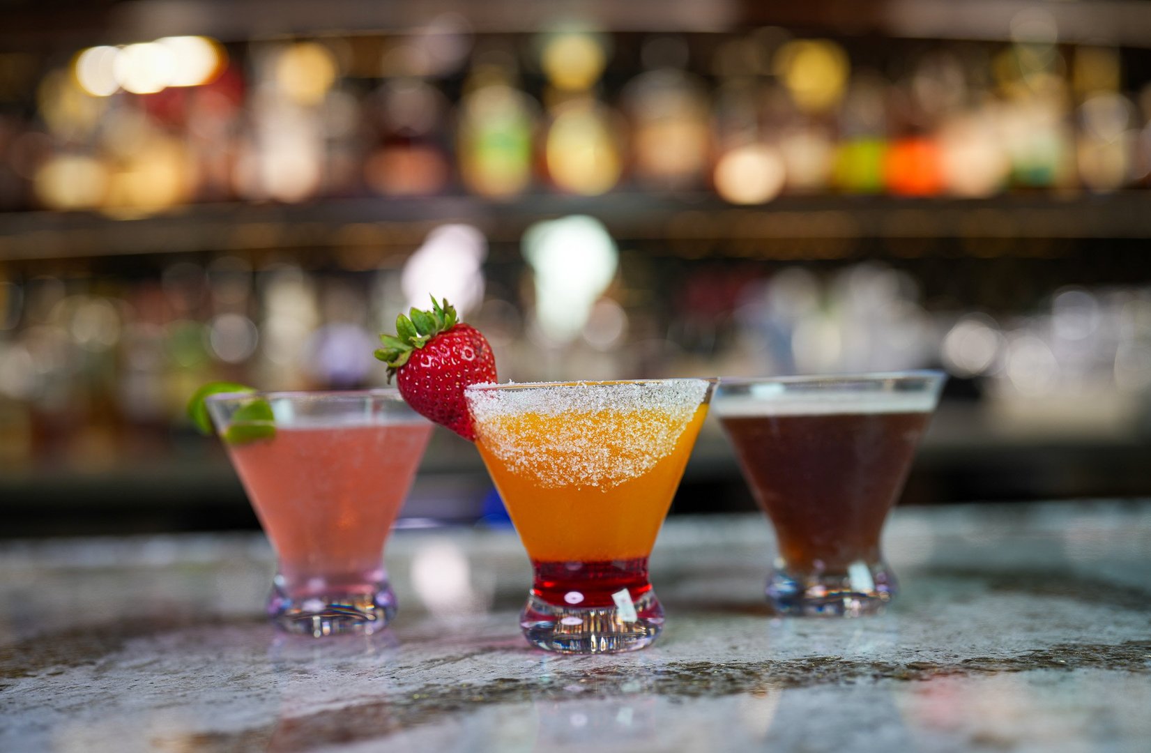 3 short martini glasses sitting on a bar. From left to right: pink flower garnished with lime peel, strawberry mango lemon drop garnished with a bright red strawberry, espresso martini