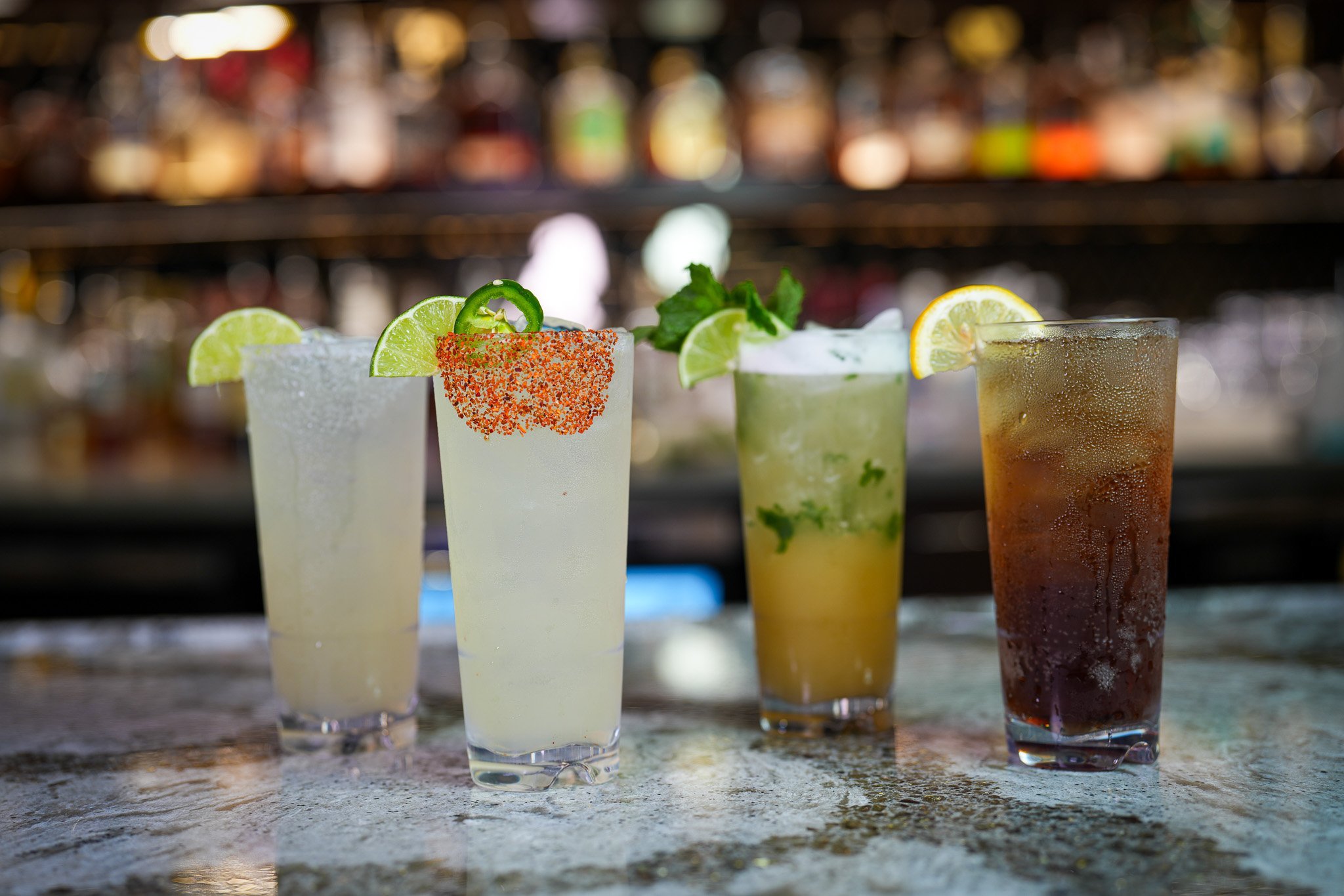 4 tall glasses of cocktails. Each looks slightly different. Left to right: cinematic margarita garnished with lime, spicy marg garnished with lime & jalapeno (tajin rim), mango cocktail with mint mixed in garnished with mint & lime, long island iced tea garnished with lemon