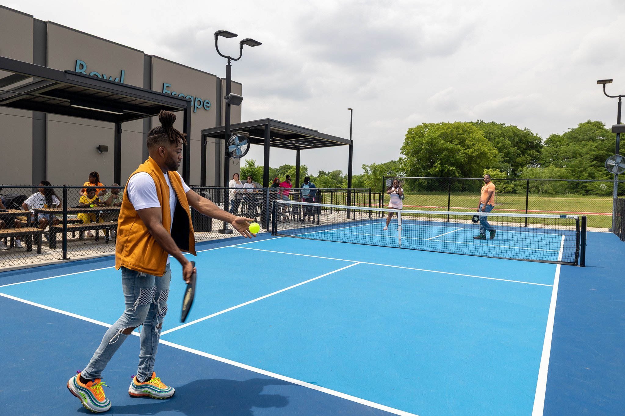 man standing in foreground getting ready to serve the pickleball. 2 players standing in the background across the court awaiting the serve.