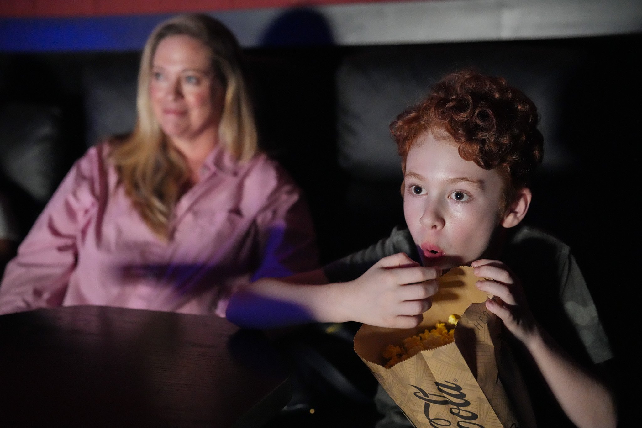 mother and child sitting in auditorium watching movie. Son is excited and eating handful of popcorn