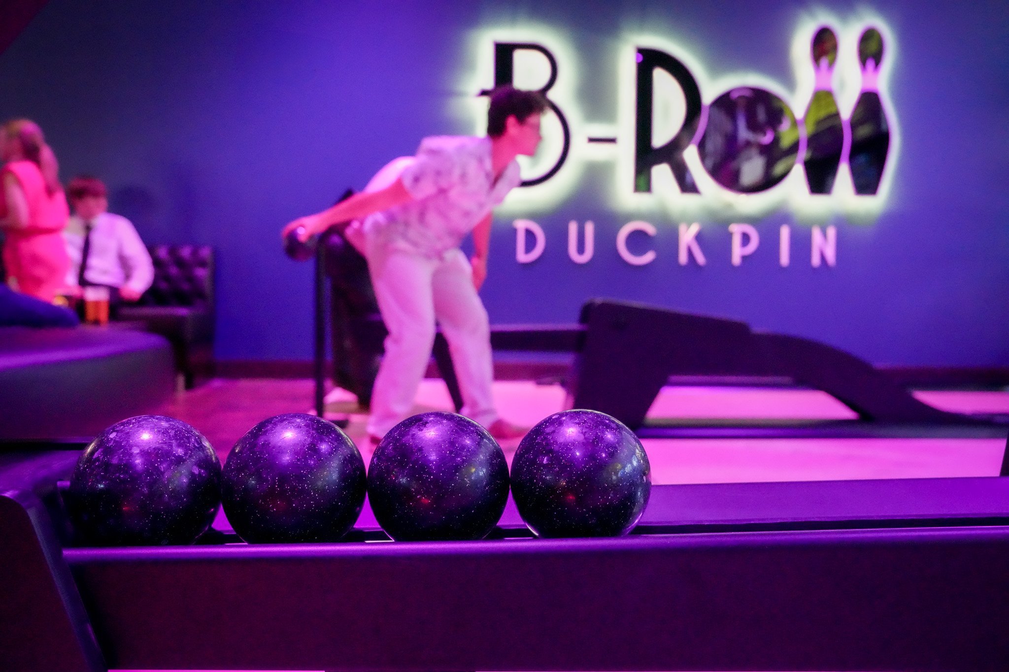 ball return holding 4 small duckpin balls. In the background is a woman with her arm back about to roll the ball. Behind her is a wall with a big sign that reads B-ROLL DUCKPIN