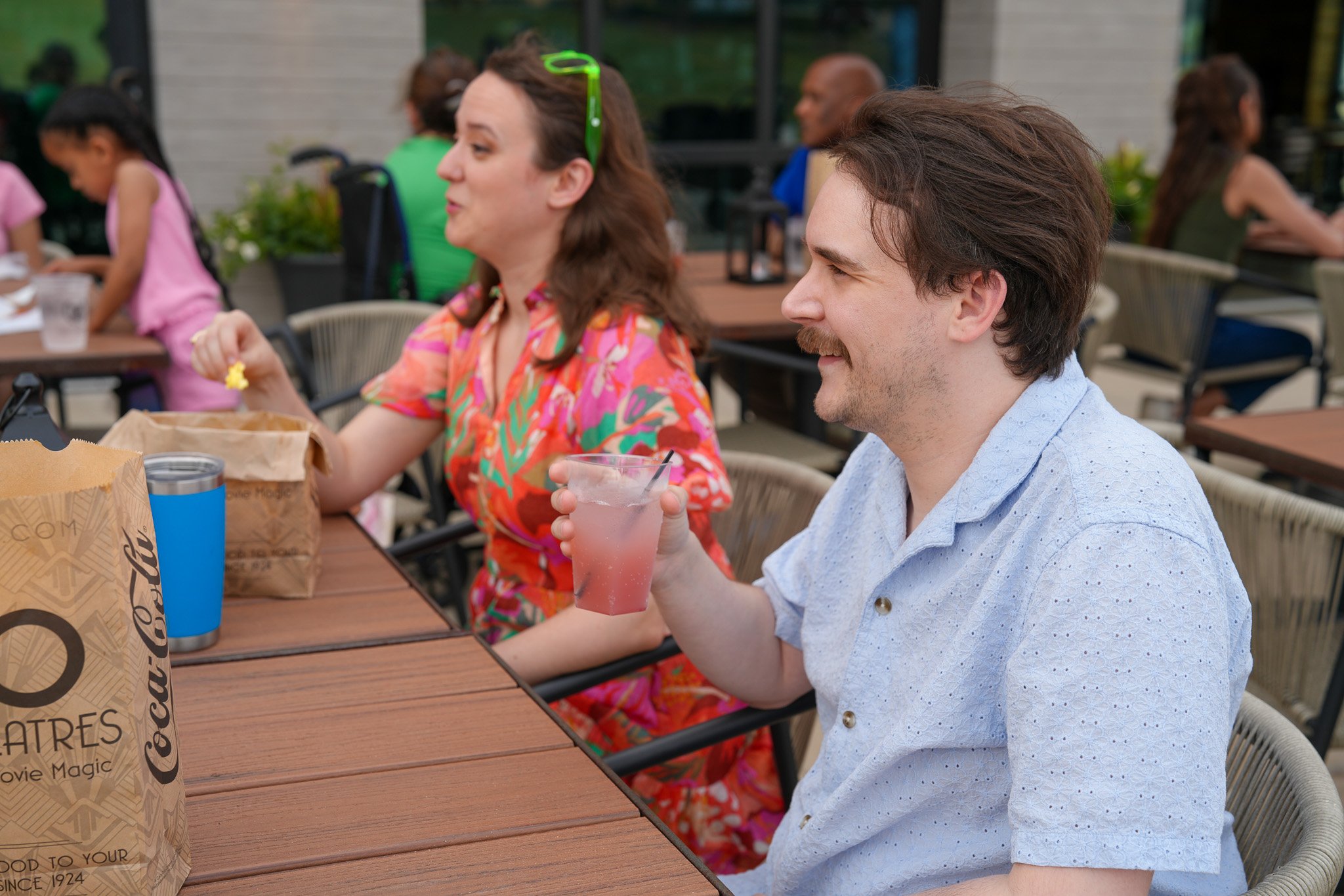 in the foreground: man and woman sitting at table outside. Drink in the man's hand, popcorn in the woman's hand. in background: other groups of people at tables.