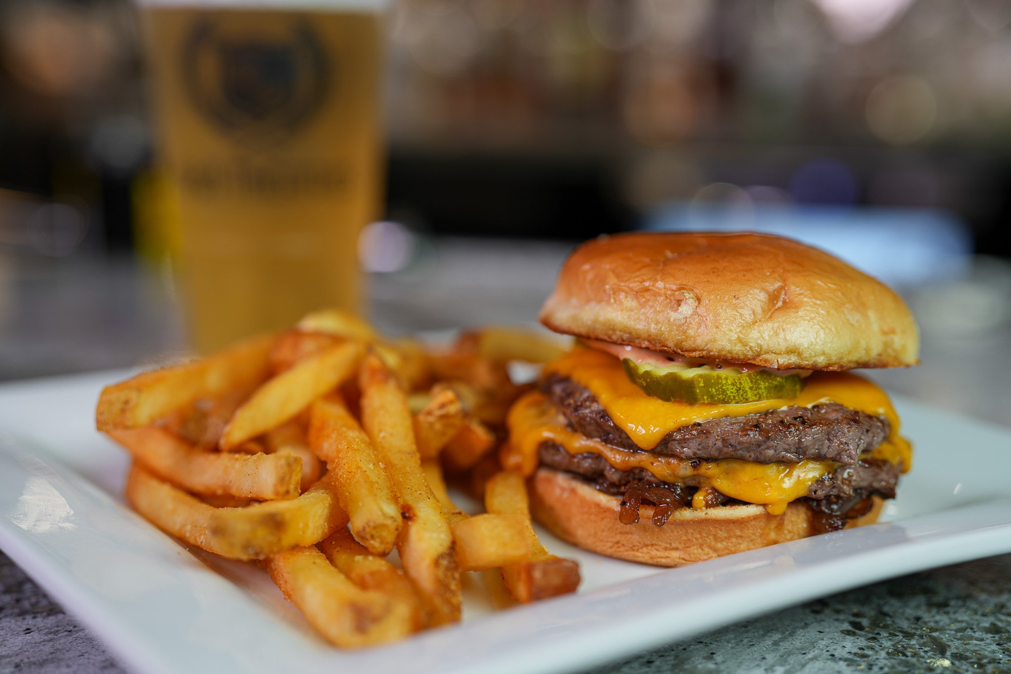 white plate filled with a double cheeseburger with pickles and special sauce plus a side of fries. Large light beer sitting behind the plate of food