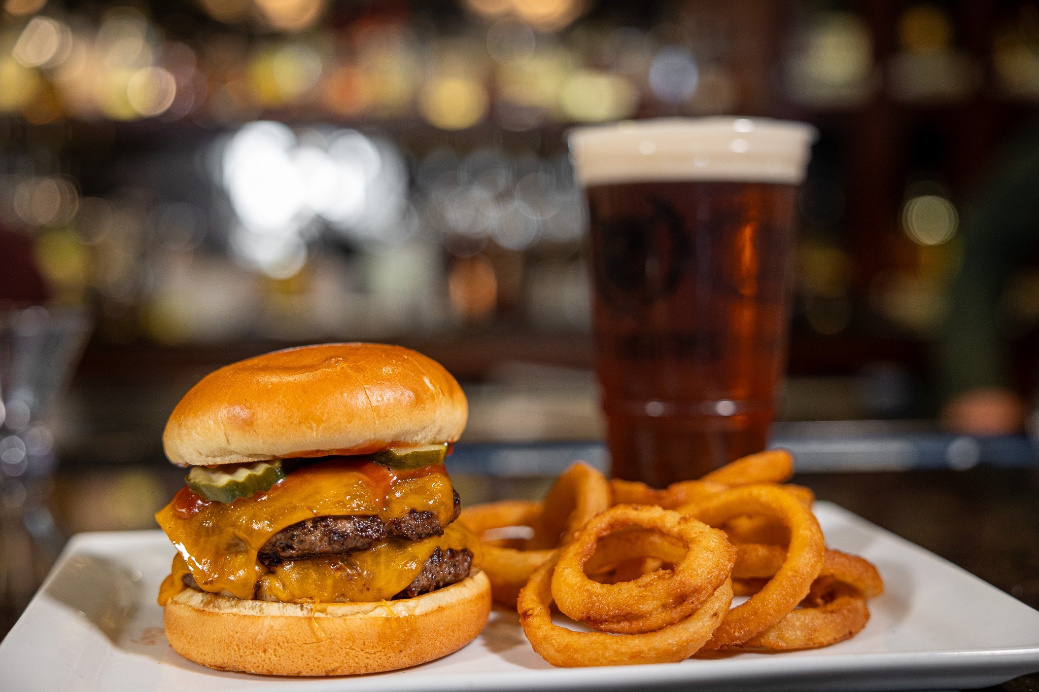 double cheeseburger with onion rings sitting on a white plate. large dark amber beer in the background