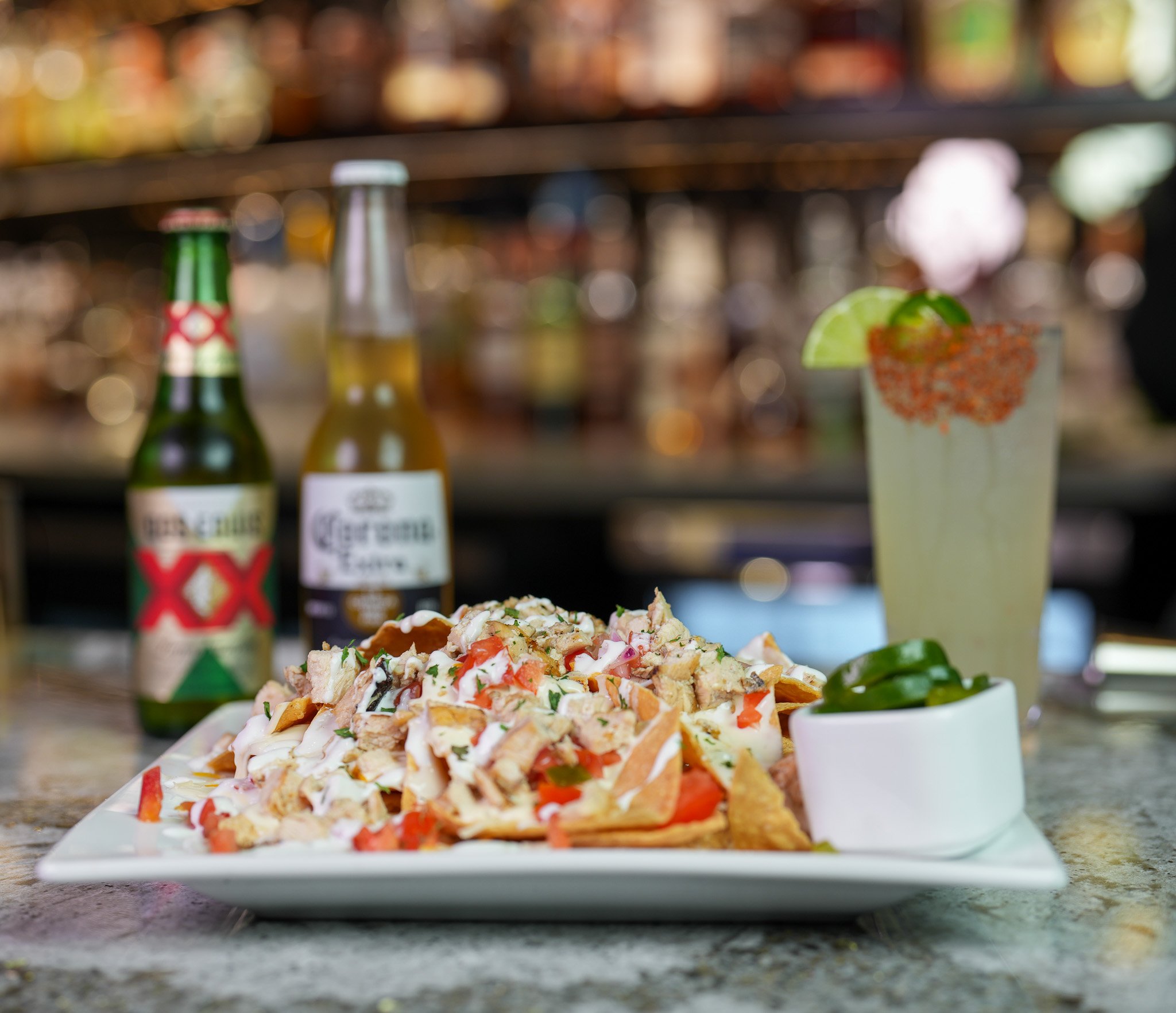 loaded chicken nachos on a white plate with a small container filled with jalapenos. In the background is a bottle of dos equis, a bottle of corona, and a glass of spicy margarita