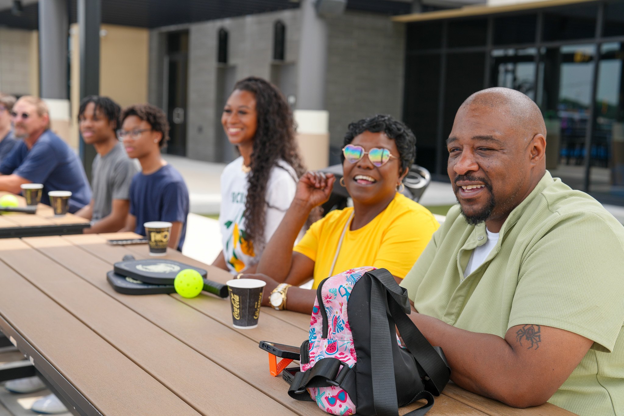 6 individuals sitting at picnic tables smiling. bags, pickleball paddles and balls, and drinks sit on the table