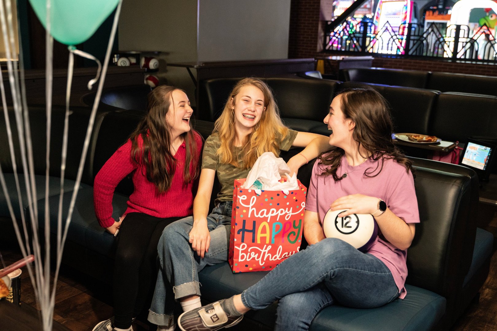 3 girls sitting on a bowling lane couch smiling and laughing with each other. between them is a big pink happy happy birthday bag with tissue paper sticking out of the top. The girl on the end on the right is holding a bowling ball with a 12 in the center