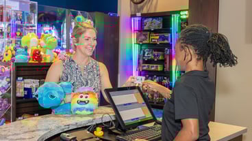 a woman is standing at a counter with stuffed alien prizes. She is smiling & handing a card to an employee. The employee's back is to the camera.