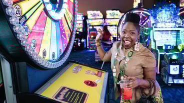 Woman is holding a cocktail & smiling towards the camera. She is holding up her right hand after she just pressed a button on a game. There is a giant wheel of fortune to the left side of the screen.