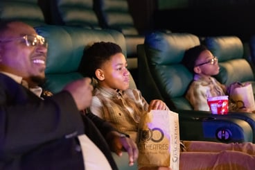 man sitting next to 2 boys. both boys are holding popcorn, the man is smiling at the screen (off screen)