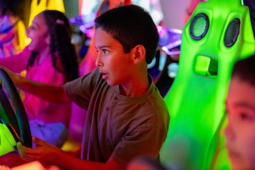 2 young boys and 1 young girl sitting in bright racing chairs. Boy in the center is holding a driving wheel & he is focused on the game