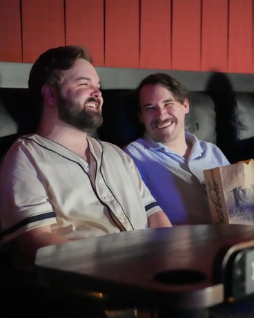 two men sitting in chairs, smiling. 1 is holding a bag of popcorn