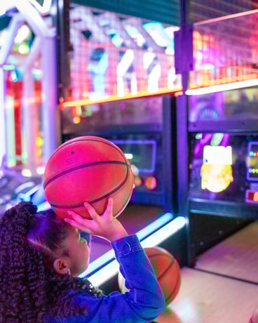 small child holding a small basketball & getting ready to shoot it into a hoop. neon lights surround the games