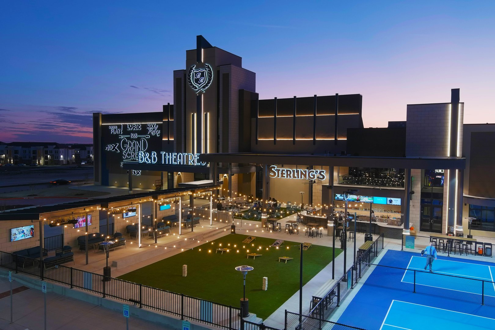 exterior drone shot of building. You can see many lit signs advertising B&B Theatres, Sterling's restaurant, and theatrical amenities. You can also see the cabanas and lawn game area, and 1 pickleball court