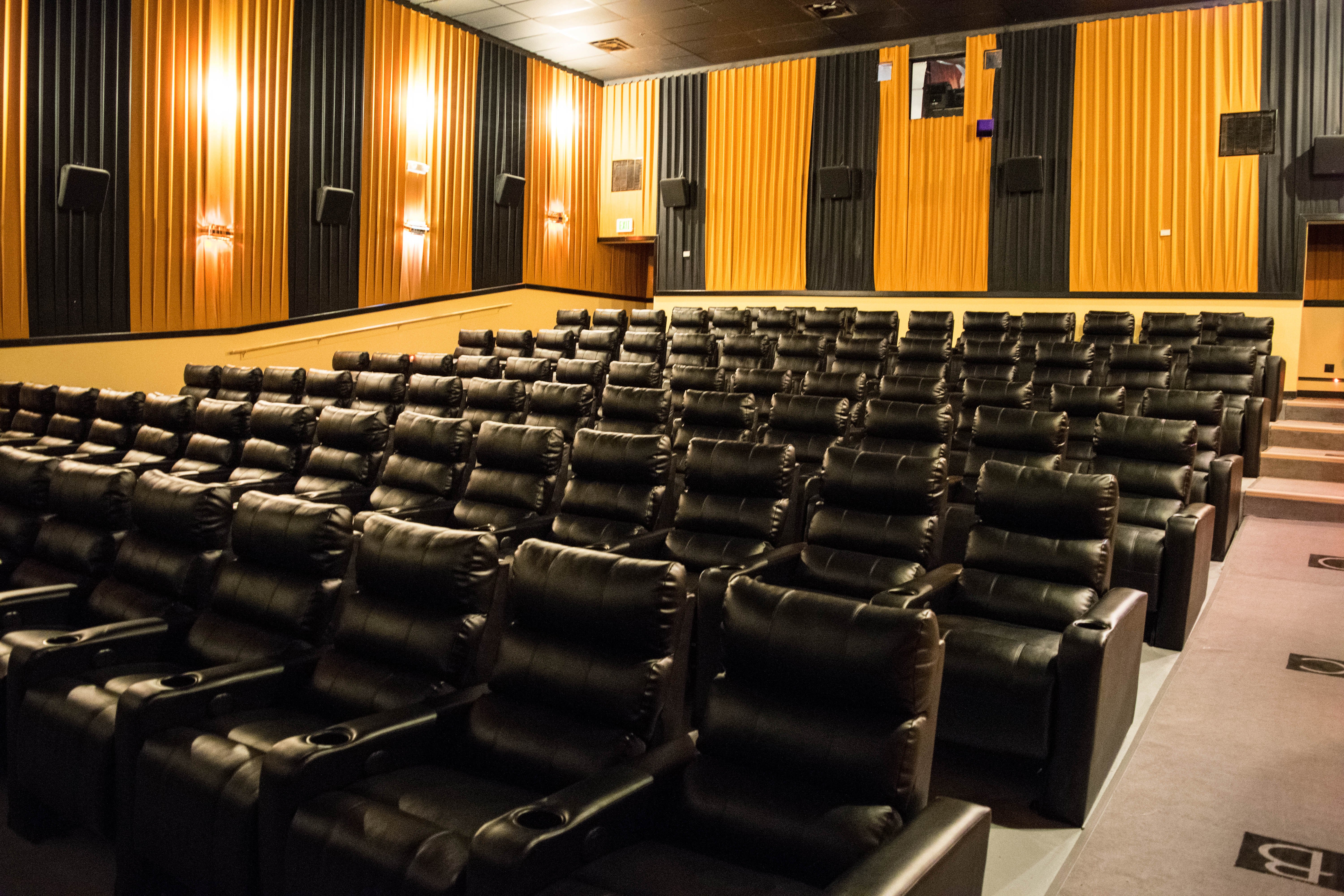 auditorium full of black recliners. Wall drapery alternate from Black to Gold panels. Lights are on in the room 