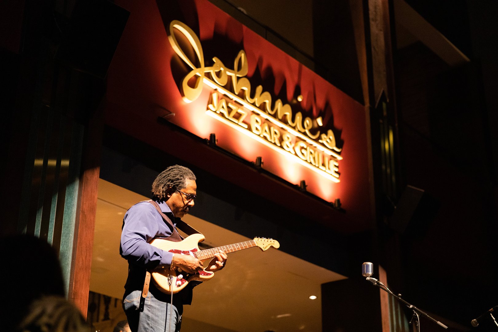man standing with guitar on stage. He is looking at guitar. Above & behind him is a sign with lights pointing on it that reads Johnnie's Jazz Bar & Grille